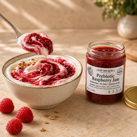 A jar of Craft Jams by Kai Prebiotic Raspberry Jam sits next to a bowl of creamy yogurt topped with raspberry jam, with a spoon lifting the jam and fresh raspberries on a rustic beige surface.