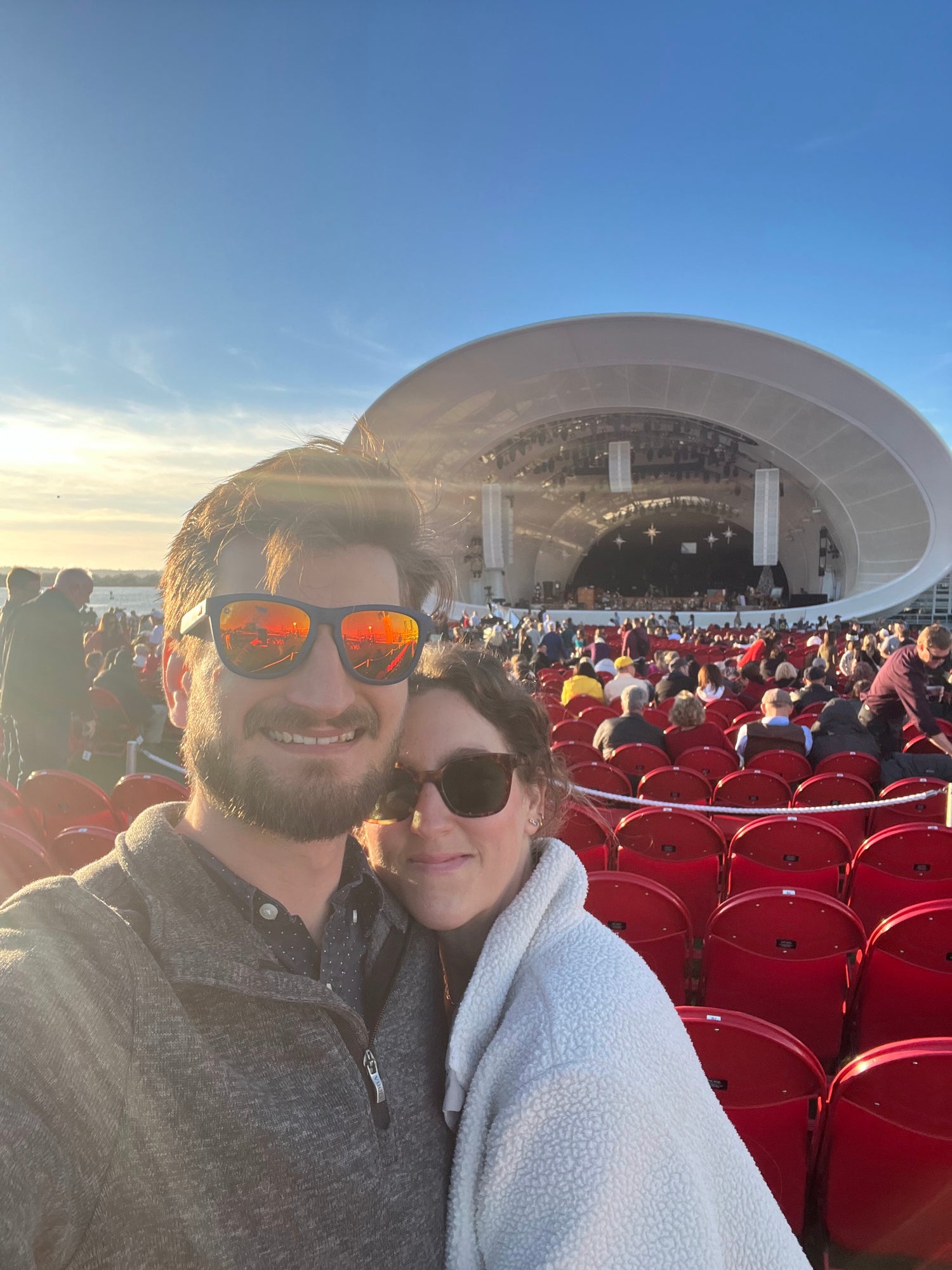 Kai and Bryon taking a selfie in front of a large outdoor amphitheater filled with people under a clear blue sky. The man wears orange mirrored sunglasses and a gray quarter-zip pullover, while the woman wears dark sunglasses and a white fleece jacket.